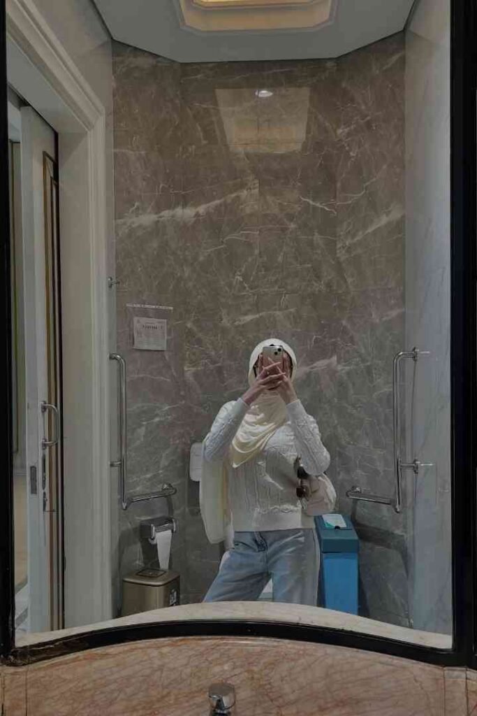 Woman in hijab and sweater taking mirror selfie in a modern bathroom